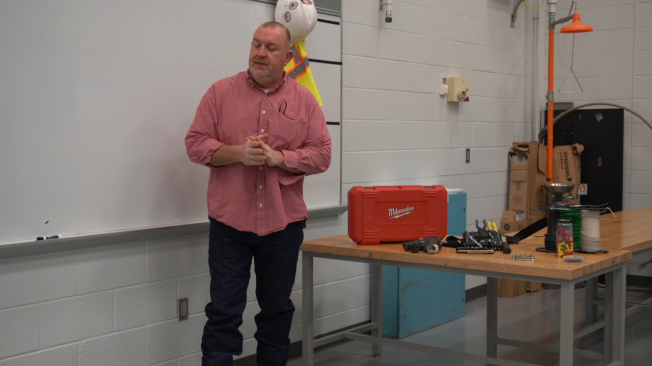 A man stands in front of a whiteboard in a classroom, with a red toolbox and various tools on a table beside him, while a yellow object hangs from the ceiling.
