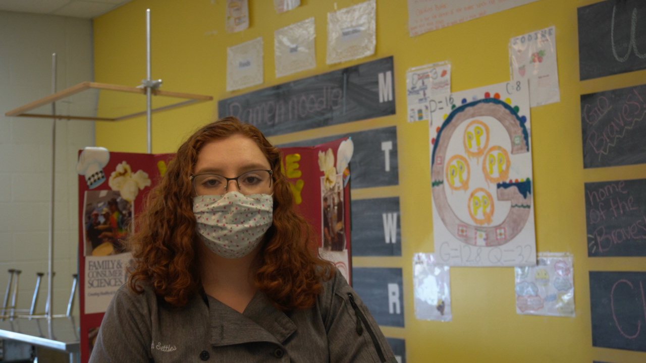 A person with curly red hair and glasses, wearing a mask, stands in front of a yellow wall decorated with educational posters and charts.