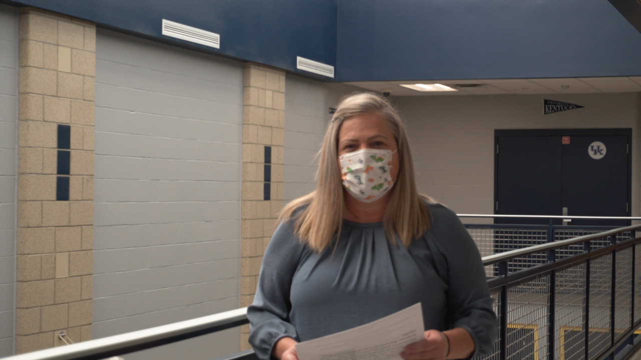 A woman wearing a patterned mask stands in a hallway, holding a piece of paper, with a blue and beige wall in the background.