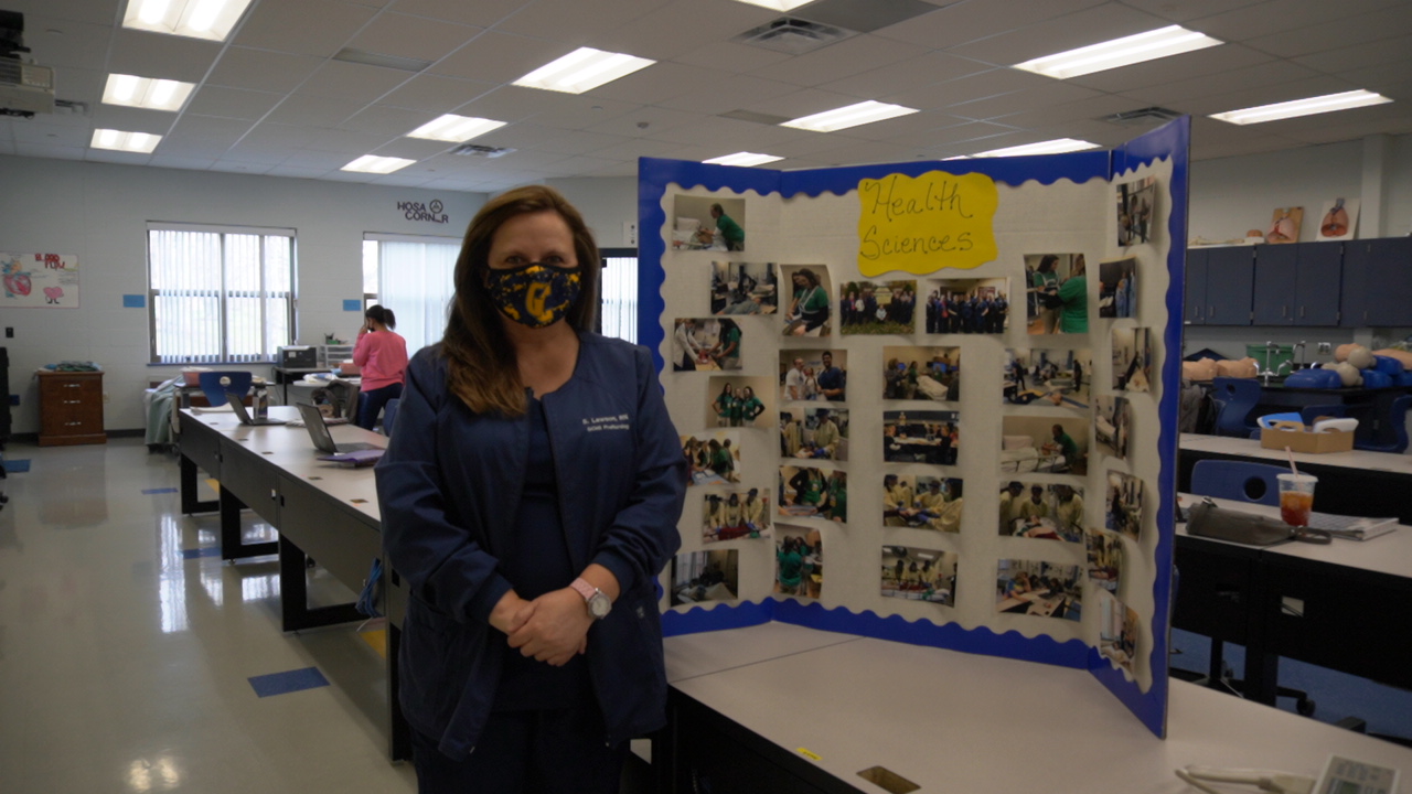 A woman wearing a mask stands beside a display board filled with photographs in a classroom setting.