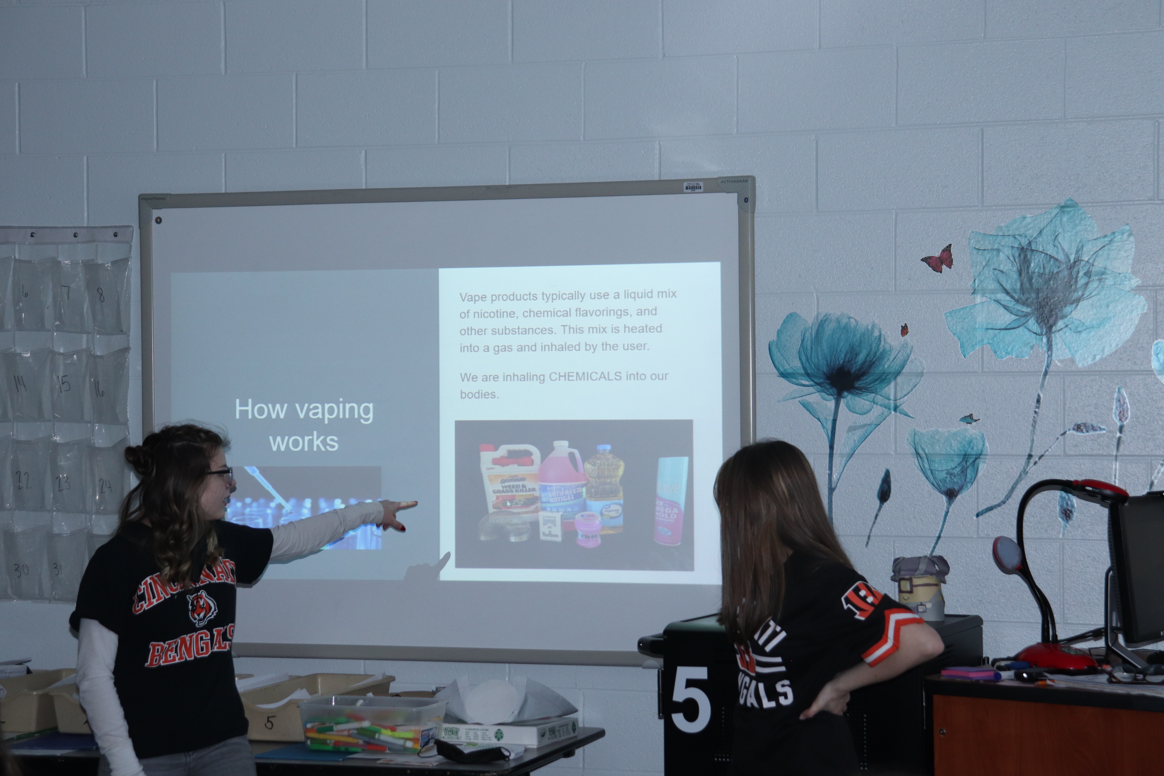 Two students are presenting in a classroom, pointing at a projector screen that displays information about vaping, while various materials are spread out on a table in front of them.