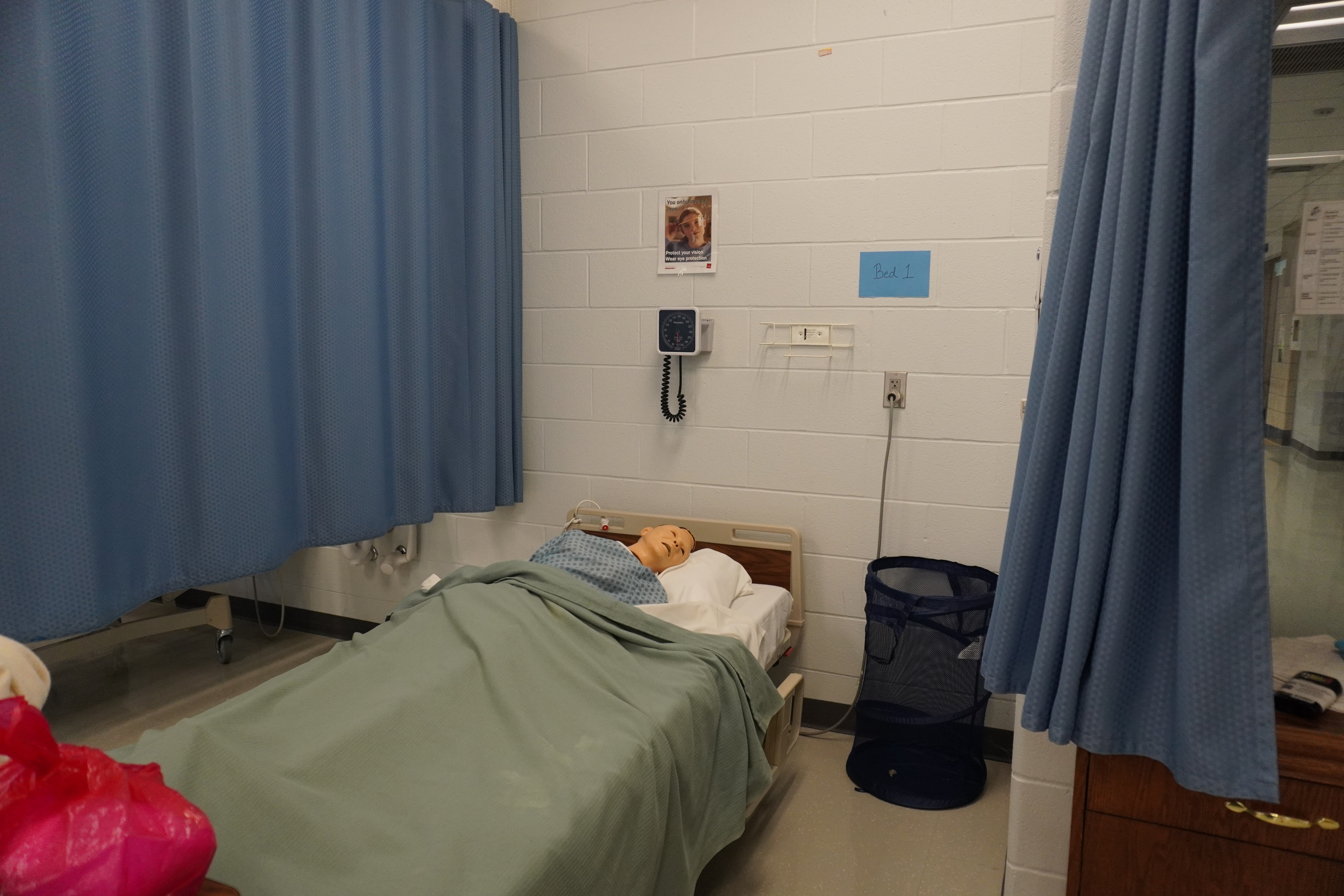 A hospital room featuring a bed with a patient mannequin, blue curtains, a medical monitor on the wall, and a laundry basket.