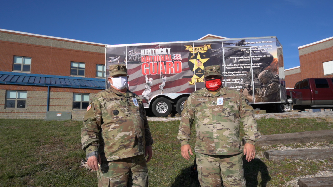 Two military personnel in camouflage uniforms and masks stand in front of a trailer displaying patriotic graphics and text, with a school building in the background.