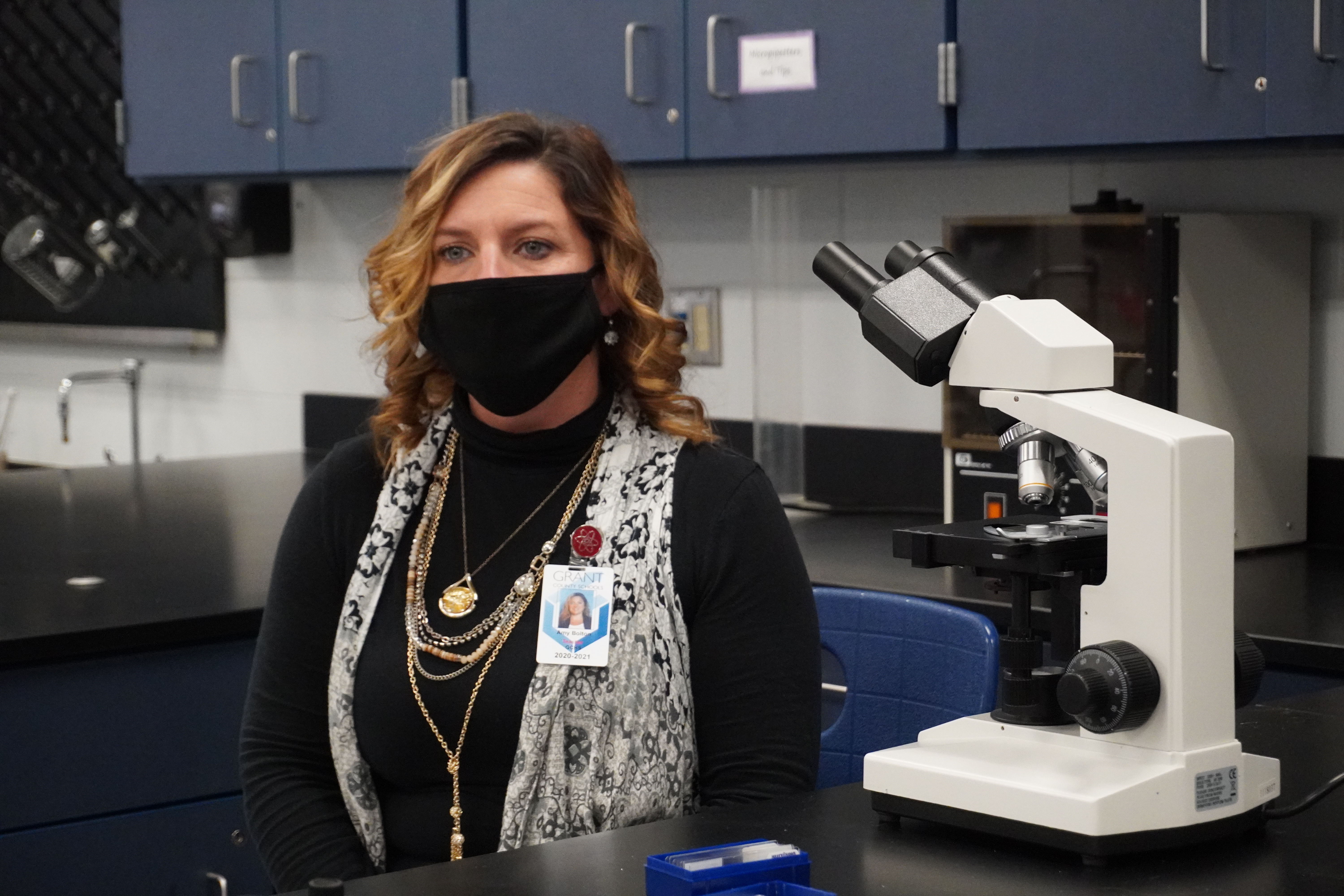 A woman wearing a black mask and a necklace sits in front of a microscope in a laboratory setting, with blue cabinets in the background.