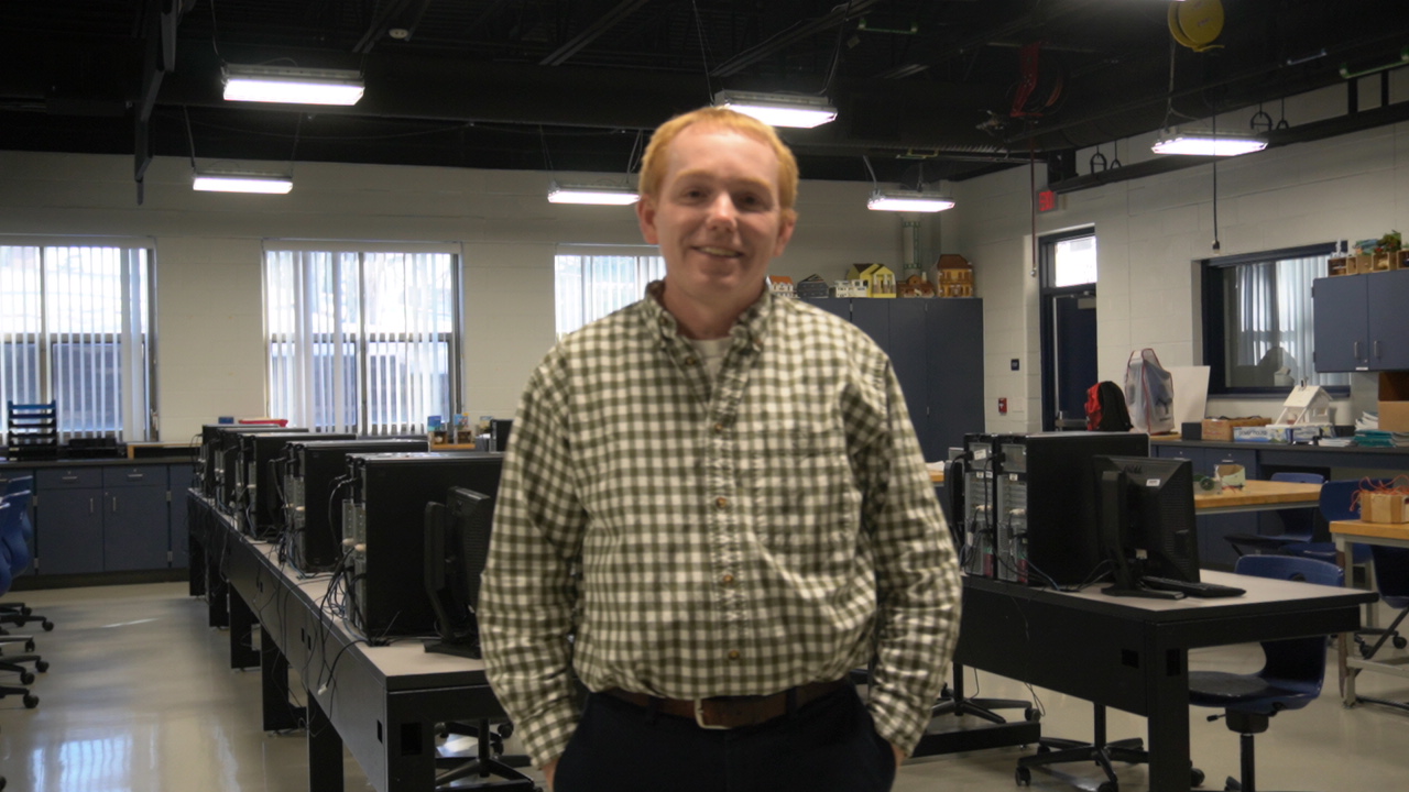 A man with short hair and a plaid shirt stands in a classroom filled with computer desks and equipment, smiling at the camera.