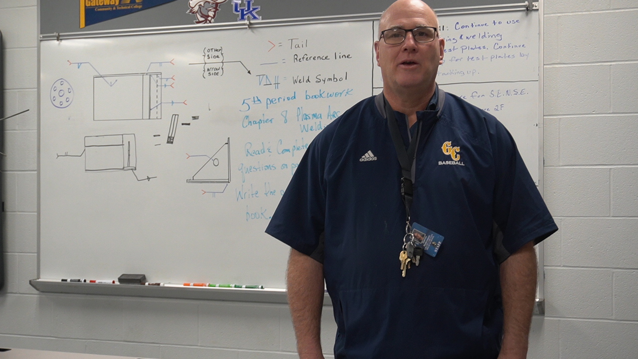 A teacher stands in front of a whiteboard filled with diagrams and notes, wearing a dark blue shirt with a school logo.