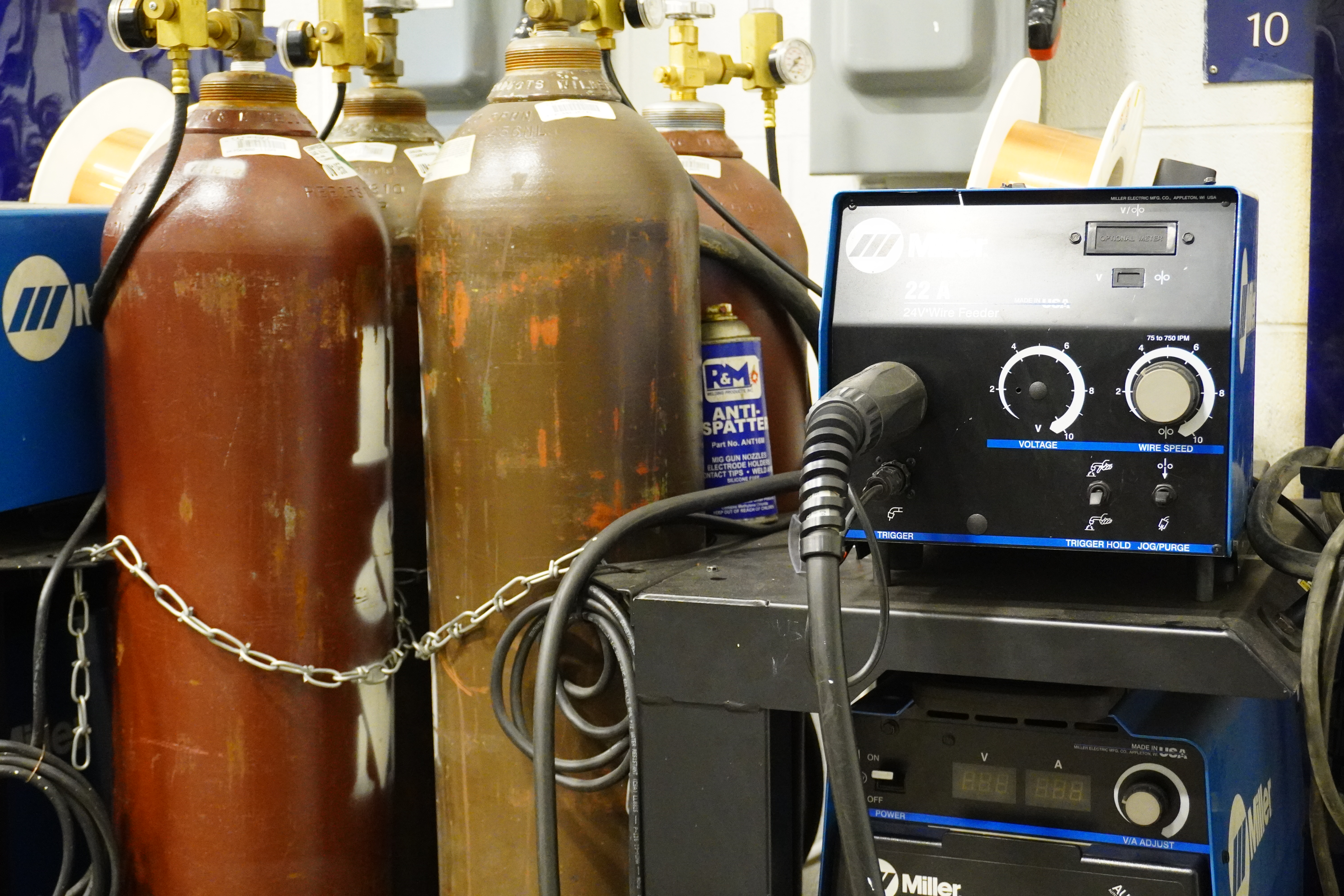 A close-up view of two gas cylinders connected by a chain, alongside a welding machine with dials and a connected hose, set in a workshop environment.