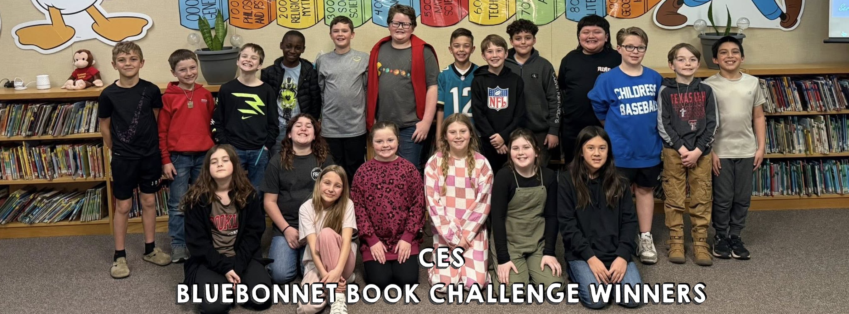A group of 20 smiling children poses in a library, standing and sitting. Bookshelves line the wall, and colorful banners hang above. Text reads "CES Bluebonnet Book Challenge Winners."