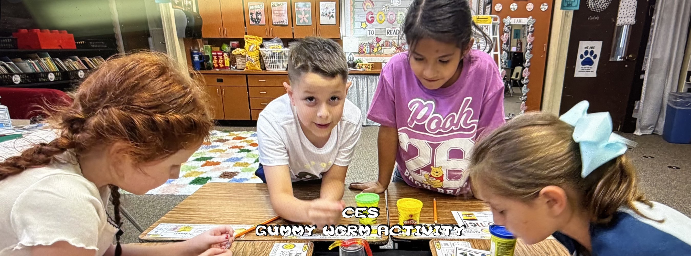 Four children gather around a classroom table for a playful gummy worm activity, sorting colorful candy pieces with focused, cheerful expressions.