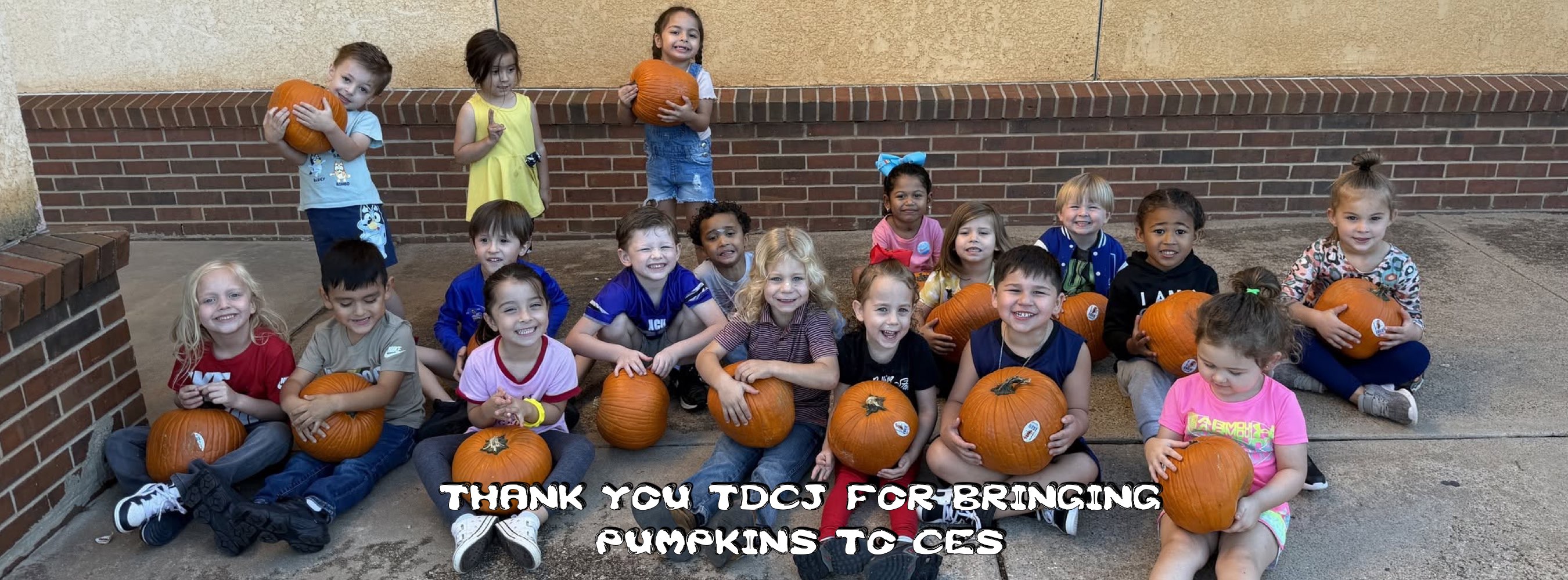 A diverse group of children sits and stands with pumpkins outside a brick building. A caption reads, “Thank you teachers for bringing pumpkins to class,” conveying cheerful gratitude.