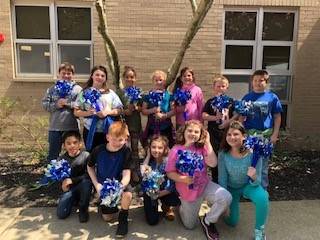 A group of eleven children, holding blue and white pom-poms, pose for a photo outside a building with large windows.