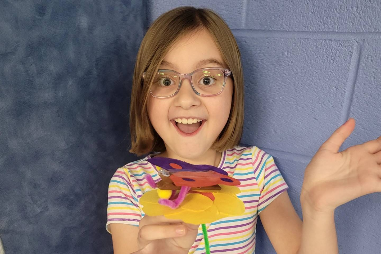 A young girl with glasses is smiling and holding a colorful craft flower made from various materials, standing in front of a blue wall.