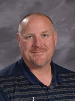 A smiling man with short hair and a beard, wearing a dark polo shirt with horizontal stripes, poses against a gray background.