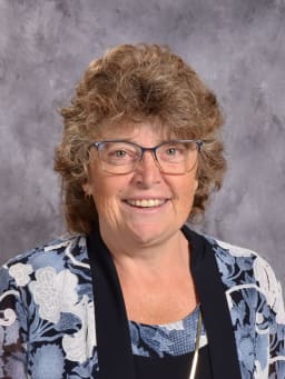 A smiling woman with curly brown hair and glasses, wearing a floral-patterned blouse, poses against a gray background.