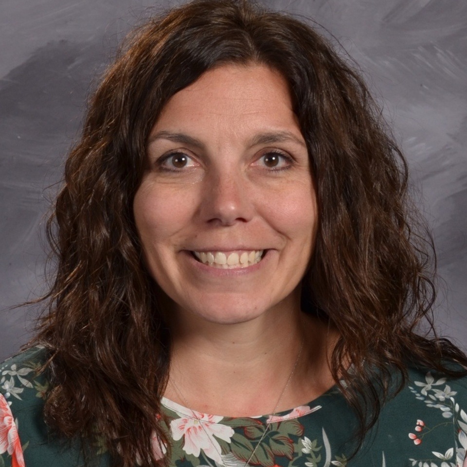 A smiling woman with curly brown hair, wearing a floral-patterned top, poses for a portrait against a gray background.
