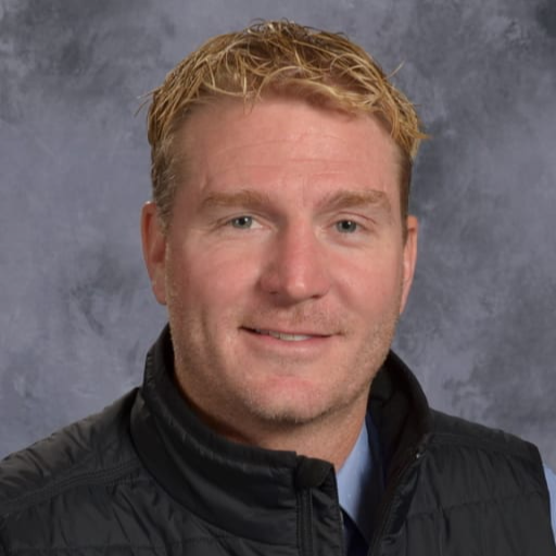 A headshot of a man with short, wavy blonde hair, wearing a black vest over a blue shirt, smiling against a gray background.