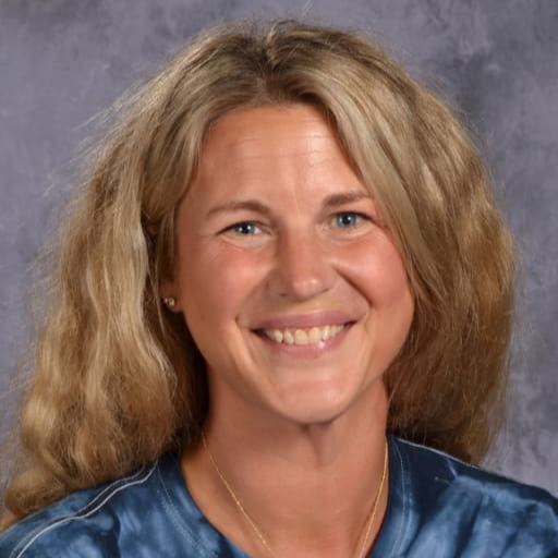 A smiling woman with long, wavy blonde hair wearing a blue tie-dye shirt, set against a gray background.