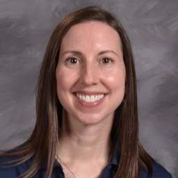 A smiling woman with long brown hair, wearing a navy blue shirt with yellow lettering, poses for a portrait against a gray background.