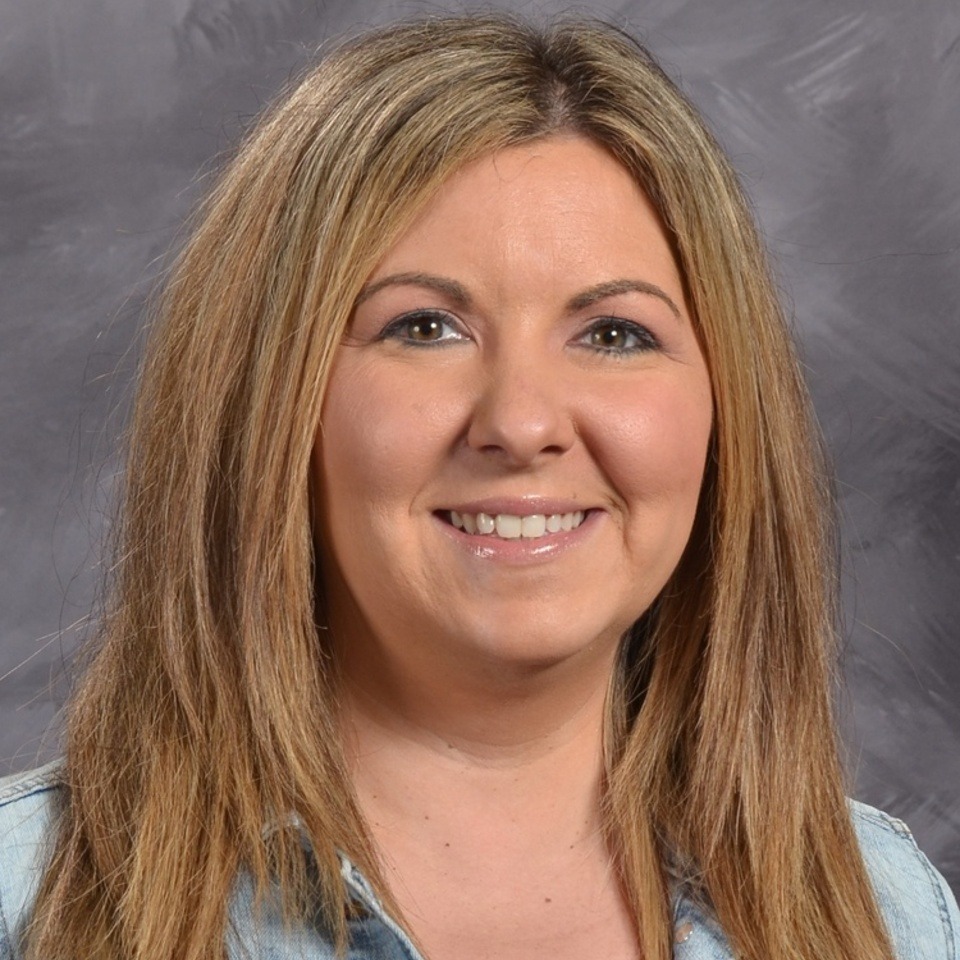 A smiling woman with long, blonde hair wearing a blue shirt, set against a gray background.