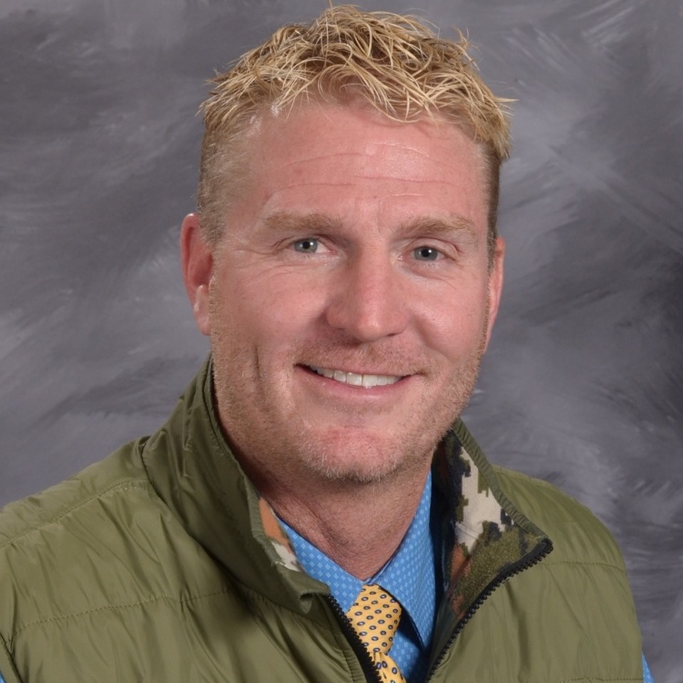 A headshot of a man with short, wavy blonde hair, wearing a green vest over a blue shirt, smiling against a gray background.