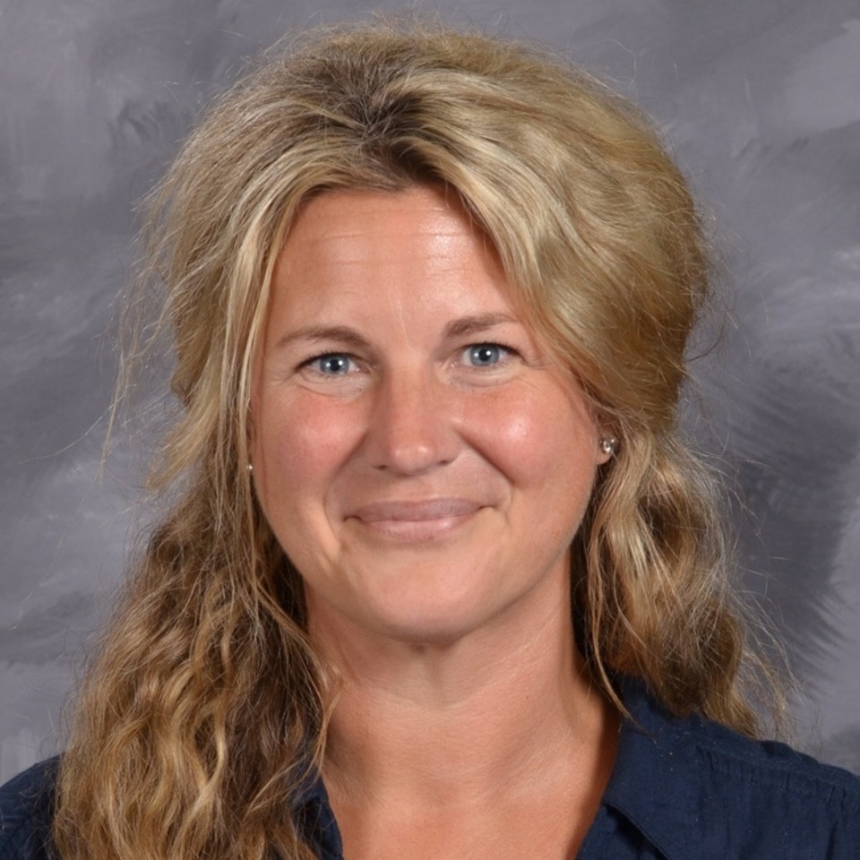 A smiling woman with long, wavy blonde hair wearing a blue shirt, set against a gray background.