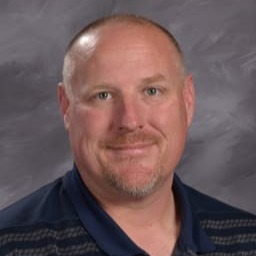 A smiling man with short hair and a beard, wearing a dark polo shirt with horizontal stripes, poses against a gray background.