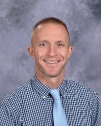 A smiling man with short hair, wearing a checkered shirt and a light blue tie, poses against a gray background.