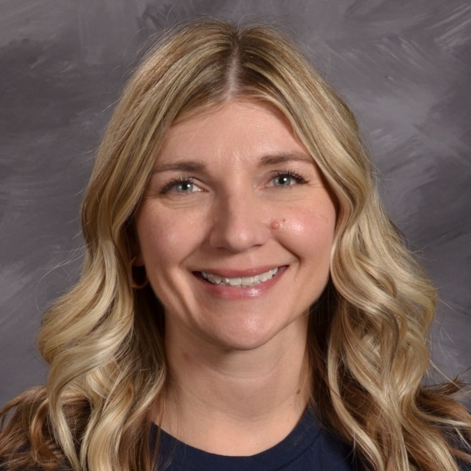 A smiling woman with long, wavy blonde hair, wearing a black top, is posed against a gray background.