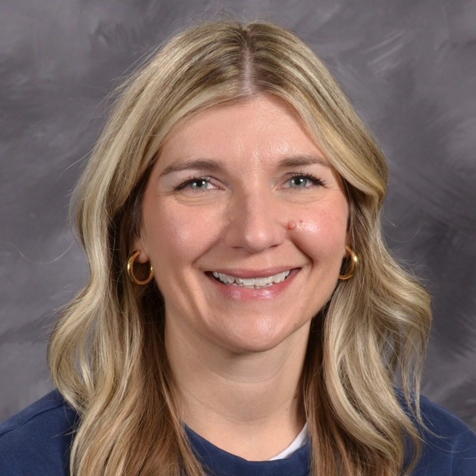 A smiling woman with long, wavy blonde hair, wearing a blue top, is posed against a gray background.