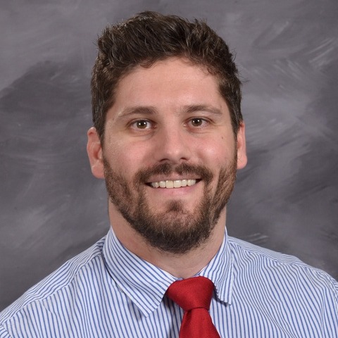 A smiling man with short, light-colored hair wearing a blue and white shirt and a red tie, set against a gray background.