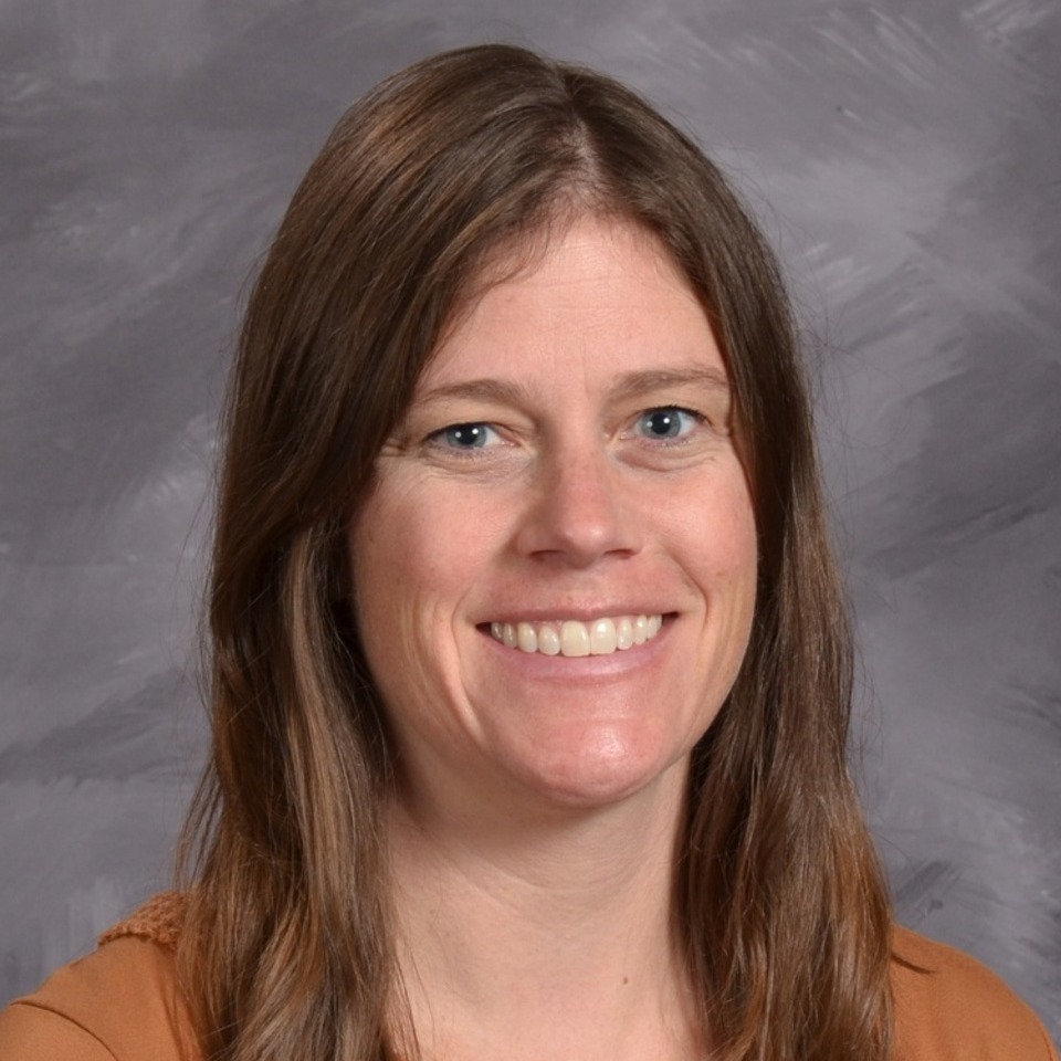 A smiling woman with long brown hair, wearing a orange shirt, is posed against a grey background.