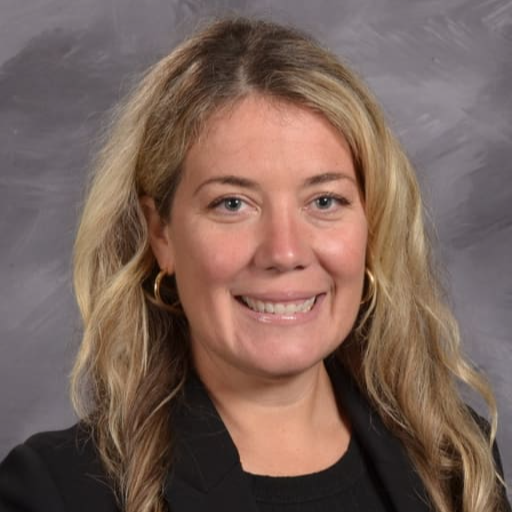 A professional headshot of a woman with long, wavy blonde hair, wearing a black blazer and smiling against a gray background.
