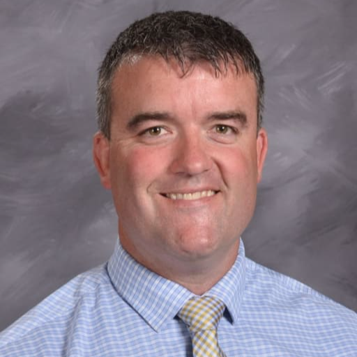 A smiling man with short, dark hair, wearing a light blue checkered shirt and a patterned tie, poses against a gray background.