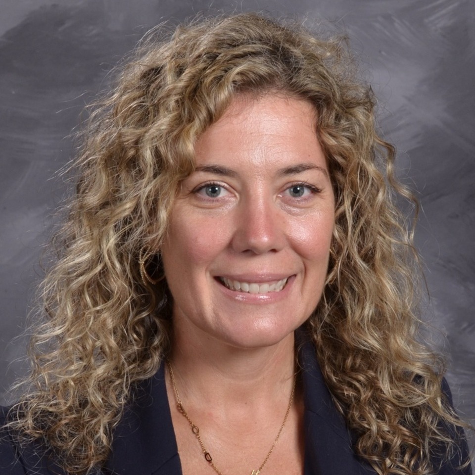 A professional headshot of a woman with long, wavy blonde hair, wearing a black blazer and smiling against a gray background.
