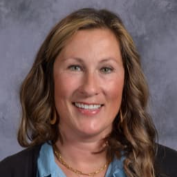 A smiling woman with wavy brown hair, wearing a blue shirt and a black cardigan, poses for a portrait against a gray background.