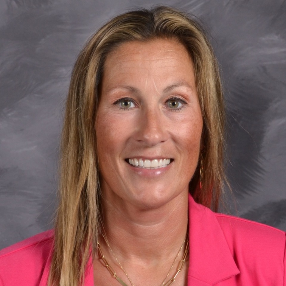 A smiling woman with brown hair, wearing a pink shirt, poses for a portrait against a gray background.