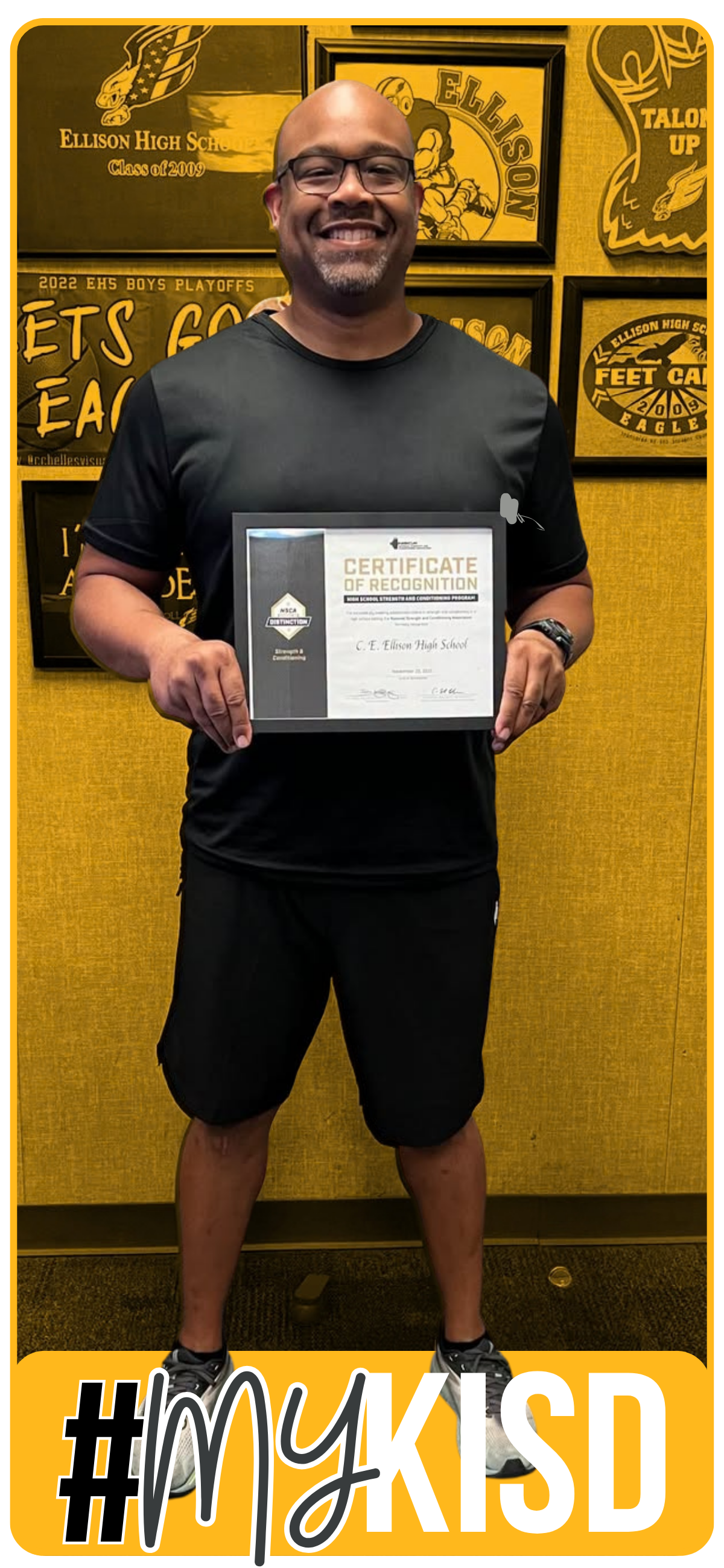 Man wearing glasses, a black shirt, and black shorts holding an award in front of a yellow background with framed plaques.
