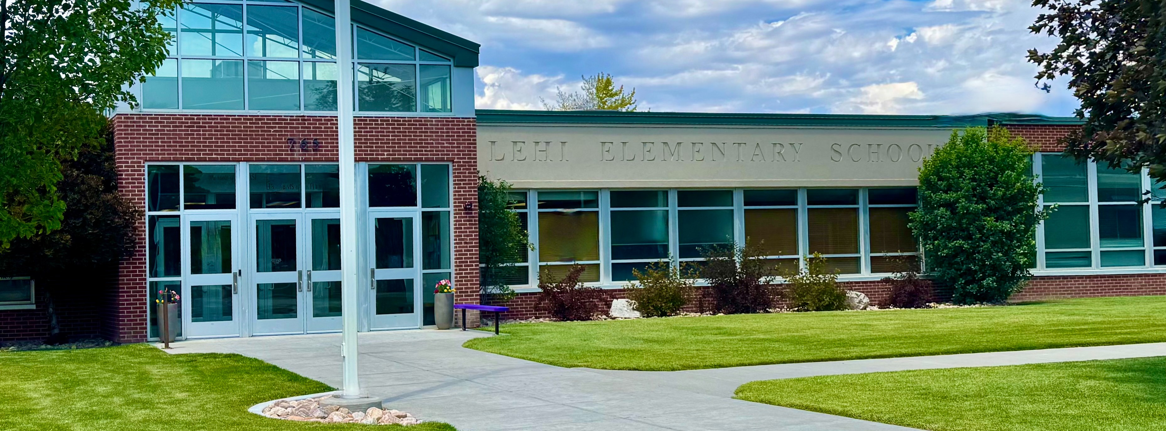 A picture of the exterior of Lehi Elementary on a summer afternoon. The grass is perfectly green and the sky is a beautiful pale blue with swashes of clouds across it. The school looks majestic and like it has seen many changes over the years, but is still strong and ready to do its job well. 