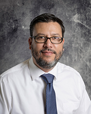 A man wearing glasses and a tie stands confidently in front of a plain wall.