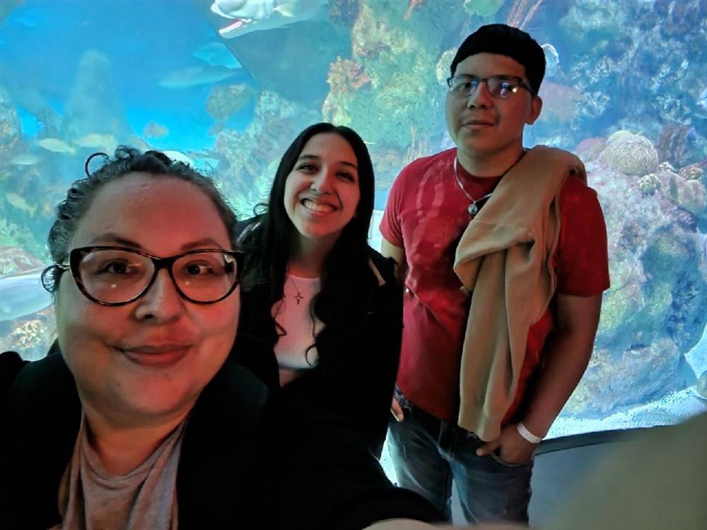 Three people pose for a selfie in front of a large aquarium filled with colorful coral and fish.