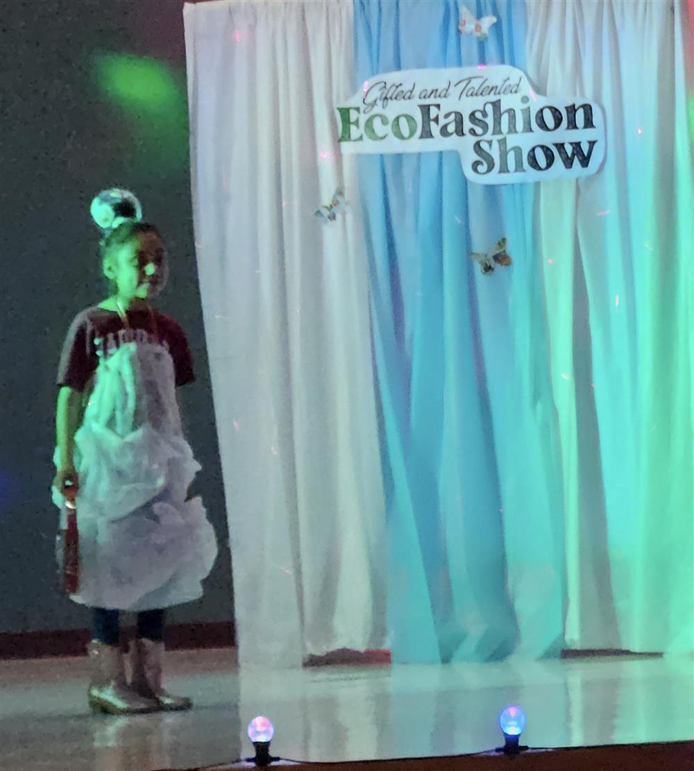 A child models a creative outfit made from recycled materials on a stage decorated with a backdrop for an EcoFashion Show.