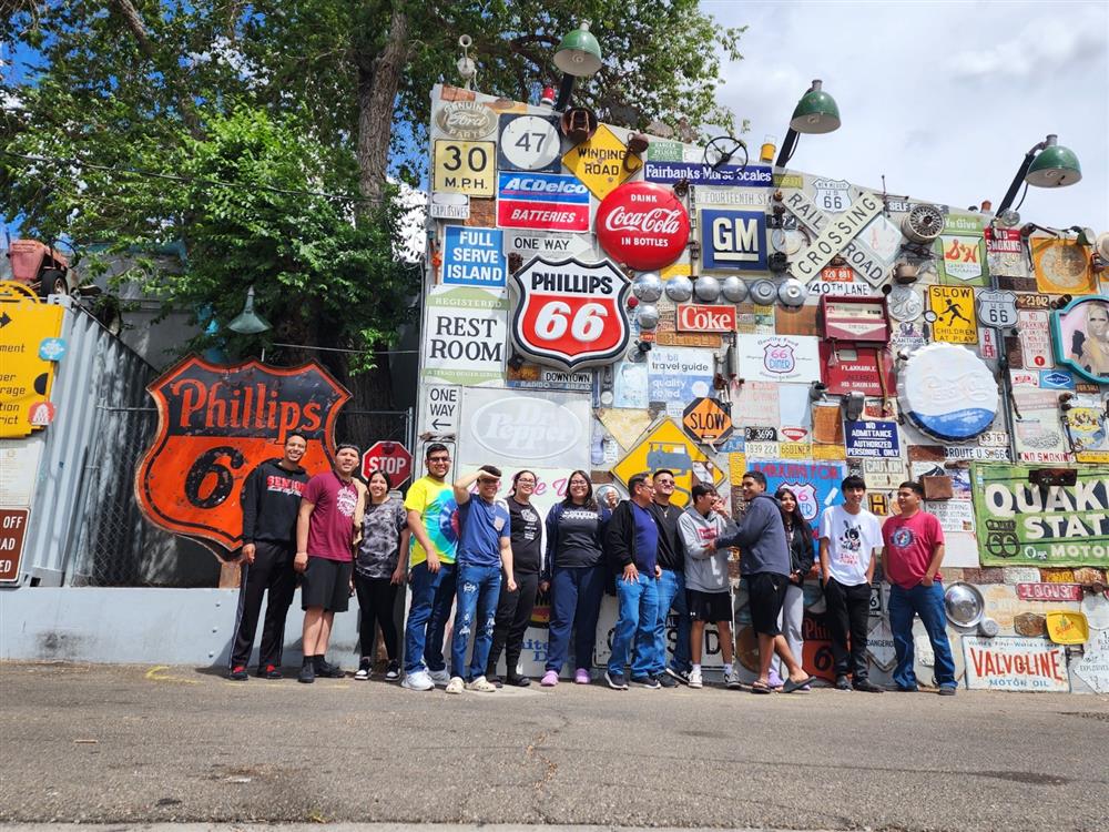 A group of people stands in front of a colorful wall covered with vintage signs and advertisements, including a prominent Phillips 66 sign.