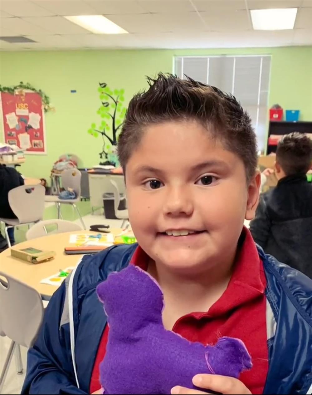 A young boy with short hair smiles while holding a purple felt shape in a classroom setting, with other students and classroom materials visible in the background.