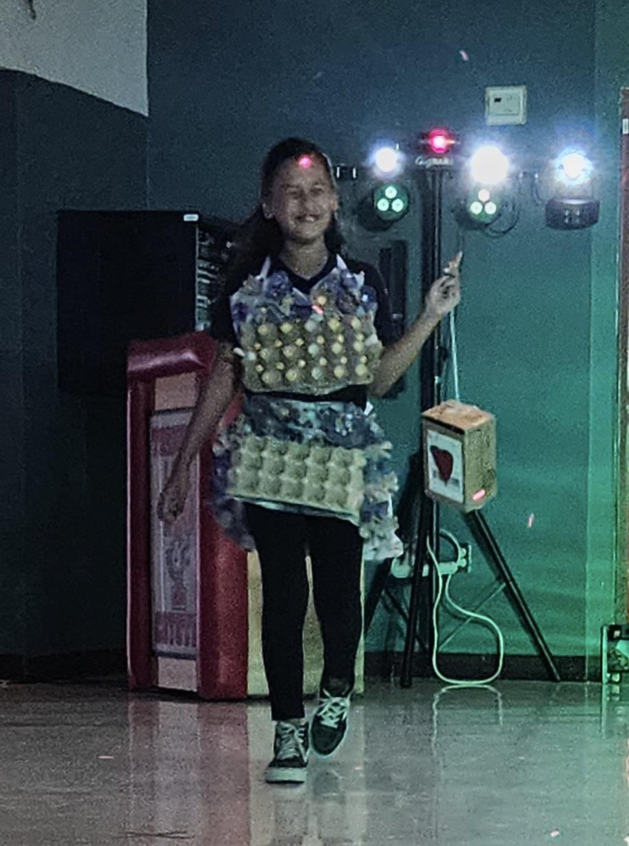 A young girl walks confidently on stage wearing a creative outfit made of egg cartons, with colorful lights and a sound system in the background.