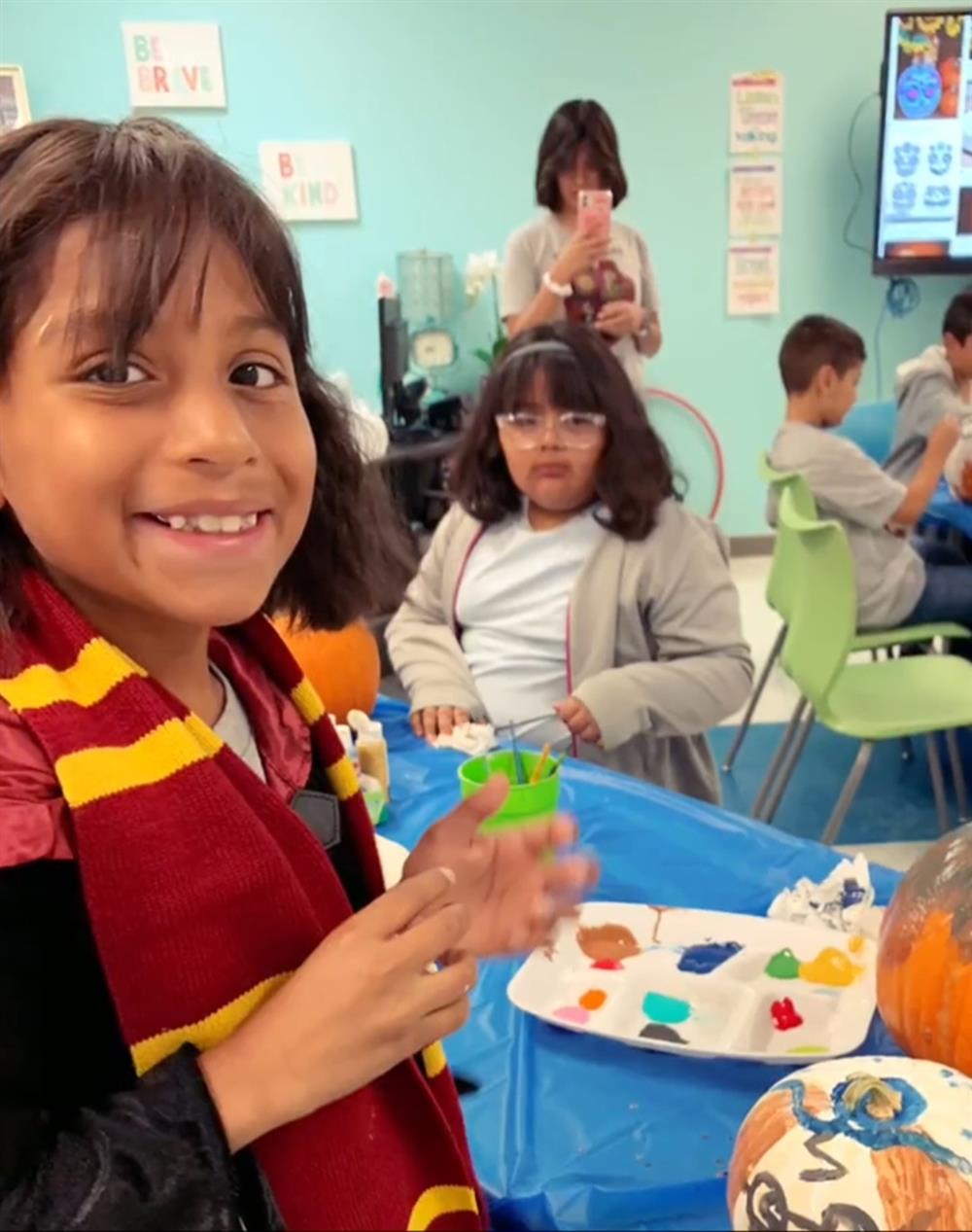 A smiling girl wearing a red and yellow scarf holds a small green cup while sitting at a table with colorful craft supplies, with other children engaged in activities in the background.