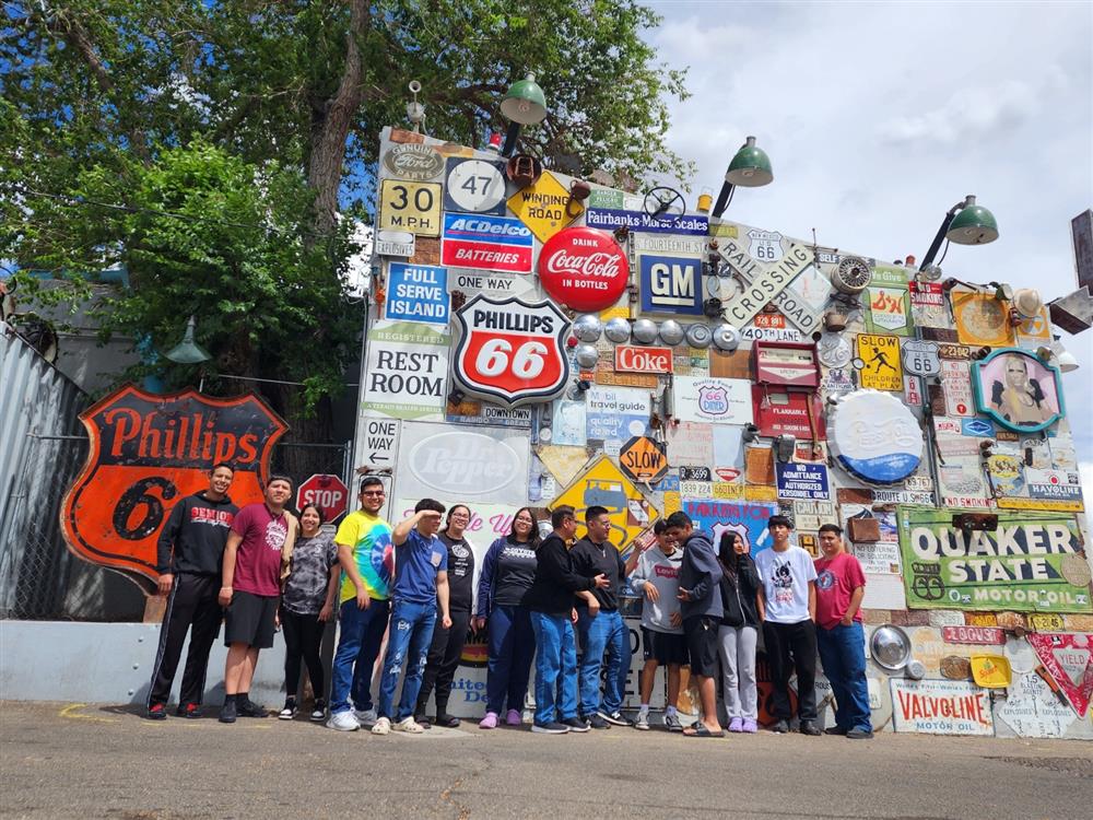 A group of people stands in front of a colorful wall covered with various vintage signs and advertisements, including a prominent Phillips 66 sign.