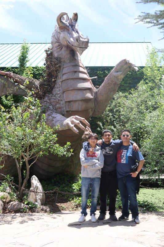 Three individuals pose together in front of a large dragon statue surrounded by greenery.