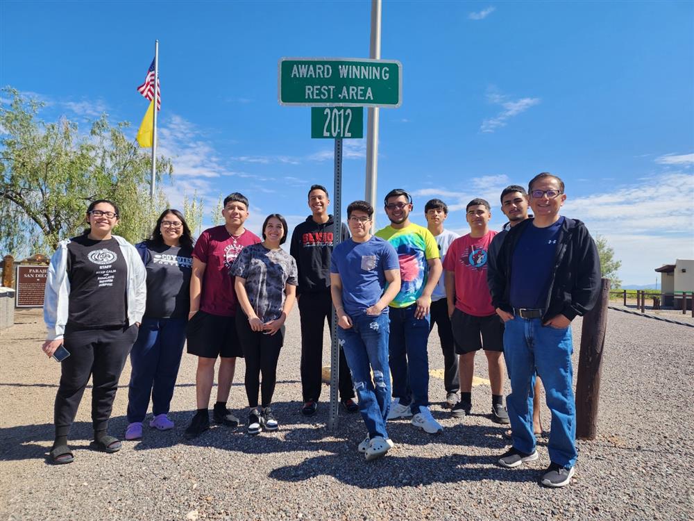 A group of eleven people stands in front of a green sign that reads 'Award Winning Best Area 2022,' with a clear blue sky in the background.