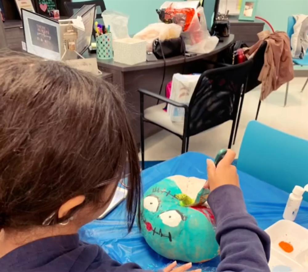 A child is painting a colorful, cartoonish rock with a face, surrounded by art supplies and a classroom setting in the background.