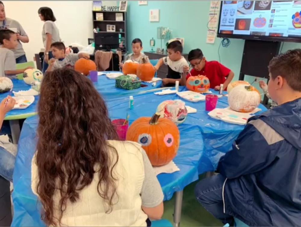 A group of children and adults are gathered around a table covered with blue plastic, painting and decorating pumpkins in a classroom setting, with a projector displaying images in the background.